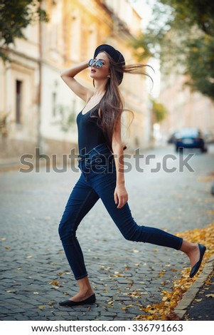 young beautiful brunette girl in a hat, sunglasses, t-shirt and jeans, with a film camera walks in the old city, holding hand hat for fear of losing it, vertical picture