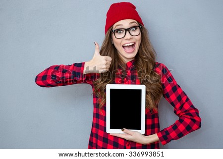 Portrait of a cheerful woman showing blank tablet computer screen and thumb up on gray background