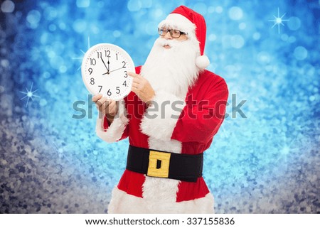 christmas, holidays, time and people concept - man in costume of santa claus with clock showing twelve pointing finger over blue glitter or lights background