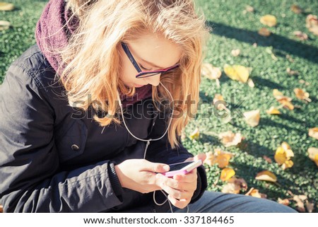 Caucasian blond teenage girl in glasses sitting in autumnal park and using smartphone. Vintage warm tonal correction photo filter, retro style effect