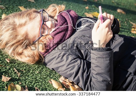 Caucasian blond teenage girl in black jacket laying in autumnal park and using smart phone. Vintage warm tonal correction photo filter, retro style effect