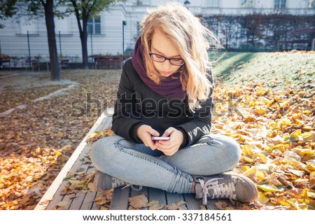 Cute Caucasian blond teenage girl in jeans and black jacket sitting on park bench and using smartphone, outdoor autumn portrait. Vintage warm tonal correction photo filter, retro style effect
