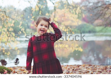 Toddler girl in red and black tartan dress playing with falling leaves and sticks standing in front of the lake holding her hair, selective focus. . Photo with applied vintage effect filters