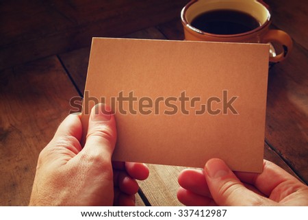 male hands holding brown empty card over wooden table background and cup of coffee. retro style image, low key and warm tones