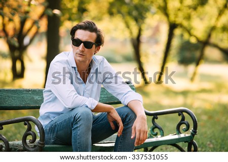 attractive man in blue shirt sitting alone on the bench in park with cellphone and sunglasses
