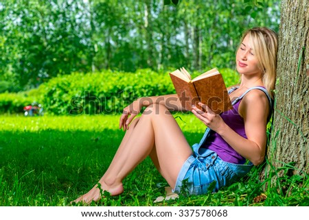 attractive young woman reading a novel in the park sitting near a tree