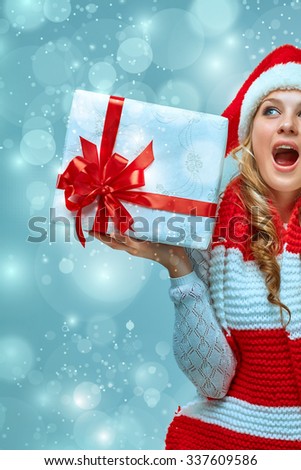Girl dressed in santa hat  with a Christmas gift. She looking at camera. Holiday concept with blue background.