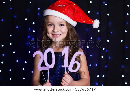 Happy child girl is wearing red santa hat holding paper digits 2016 in hands in a studio over background scene with blue lights, new year and christmas