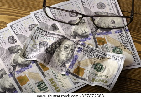 Glasses on top of the $ 100 bills and crumpled $ 100 bill on a wooden background.