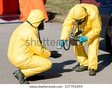 Scientist dosimetrist with radiometer and scientist radiation supervisor in protective clothing and gas masks examine a  sample of radioactive material in hazardous radioactive zone. Royalty-Free Stock Photo #337705694