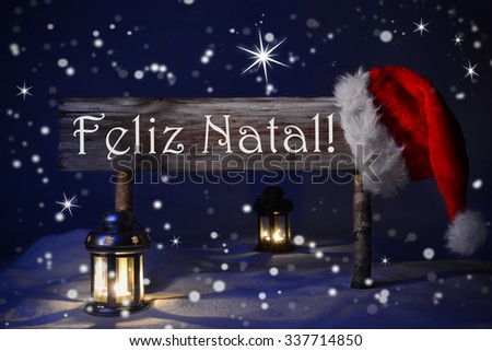 Wooden Christmas Sign And Santa Hat With Snow. Portuguese Text Feliz Natal Means Merry Christmas For Seasons Greetings. Blue Silent Night With Snowflakes And Sparkling Stars. Lantern And Candlelight