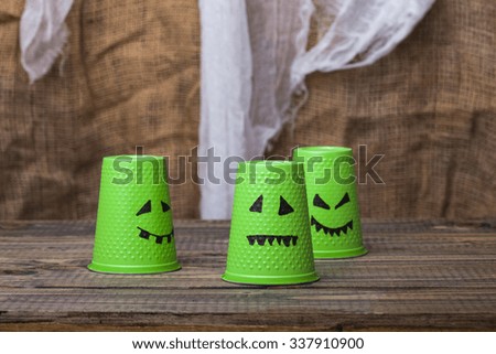 Photo set of three green disposable cups with Halloween ghost scary faces smiles with teeth drawn in black felt pen standing on wooden table over blurred rustic background, horizontal picture 