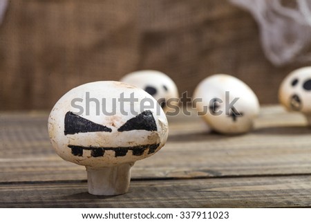 Photo closeup still life of one Halloween white champignon with ghost face smile drawn in black felt pen standing on wooden table on blurred group of button mushrooms background, horizontal picture 