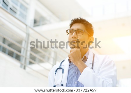 Portrait of Asian Indian medical doctor thinking and looking away, standing outside hospital building, beautiful golden sunlight at background.