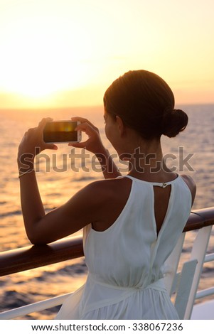Cruise ship vacation woman taking photo with smart phone camera enjoying sunset on travel at sea. Girl using smartphone to take picture of ocean sunset. Woman in dress on luxury cruise liner boat.