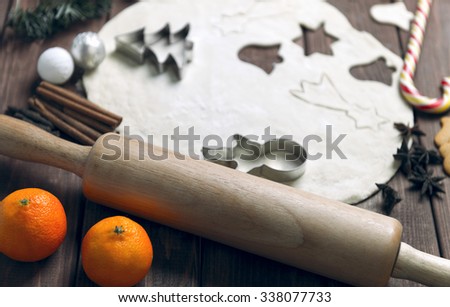 On a wooden desk background Christmas composition - candy cane, dough figurines, tree branch balls, raisins, nuts, cinnamon sticks, cookies, anise, mandarins; bump; roll out the dough layer; plunger