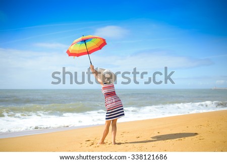 Picture of beautiful young woman with rainbow umbrella standing on seashore. Backview of pretty lady in hat on blurred summer sky outdoor background.