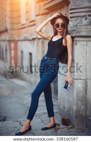 oung beautiful brunette girl in a hat, sunglasses, t-shirt and jeans with a film camera in hand walks along the ancient city