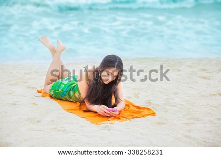 Beautiful biracial Asian Caucasian teen girl lying on tropical beach with cell phone 