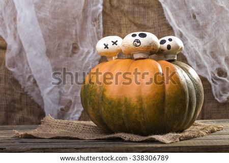 Photo still life one big whole fresh orange green pumpkin with three Halloween champignons with ghost faces drawn in black on top on sackcloth on wooden table on rustic background, horizontal picture 