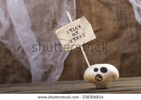 Photo closeup still life one white champignon ghost face with happy Halloween lettering trick or treat on white paper sheet on stick on wooden table on blurred rustic background, horizontal picture 
