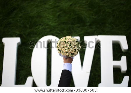 Closeup view of one beautiful fresh bright white yellow big wedding bouquet of rose flowers in human hand of bride and love text of letters lying on green grass, horizontal picture