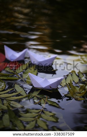 Closeup view of many beautiful colorful autumn tree leaves red yellow orange green colors floating on wavy water with reflection of nature with white paper ship on outdoor background, vertical picture