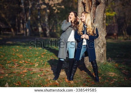 Two happy and cheerful young girl student, blond and brunette in coat and jeans laughing in sunny autumn park full of fallen leaves, horizontal pictures