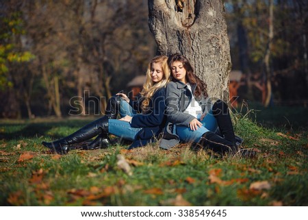 Two happy and cheerful young girl student, blond and brunette in coat and jeans laughing in sunny autumn park full of fallen leaves, horizontal pictures