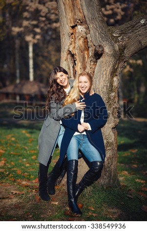 Two happy and cheerful young girl student, blond and brunette in coat and jeans laughing in sunny autumn park full of fallen leaves, vertical picture