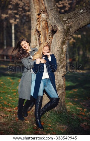 Two happy and cheerful young girl student, blond and brunette in coat and jeans laughing in sunny autumn park full of fallen leaves, vertical picture