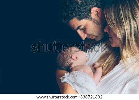 Portrait of young happy couple embracing and looking to their newborn over a dark background. Family and baby care concept. Royalty-Free Stock Photo #338721602