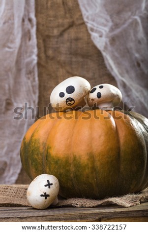 Photo still life one big whole orange green pumpkin with three Halloween champignons with ghost faces drawn in black on top and on sackcloth on wooden table on rustic background, vertical picture 