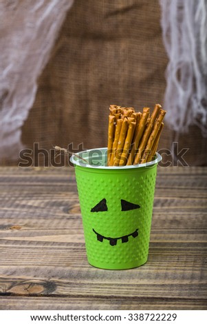 Photo still life one disposable green cup with Halloween ghost face drawn in black felt pen containing straws peaking out standing on wooden table over blurred rustic background, vertical picture 