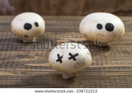 Photo still life set of three Halloween white button mushrooms champignons with ghost face eyes drawn in black felt pen standing on wooden table over blurred rustic background, horizontal picture 