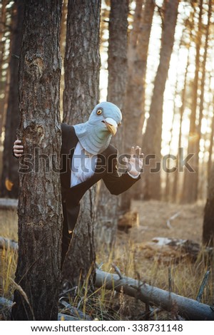 Creepy man in a suit and a rubber bird mask, standing in the sunset forest, behind the tree, waving to the camera 