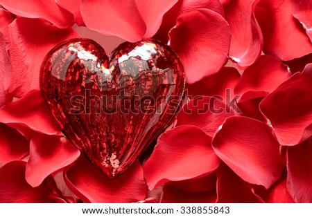 Vibrant red heart made of glass surrounded with scarlet rose petals, symbol of true love and passion, top view