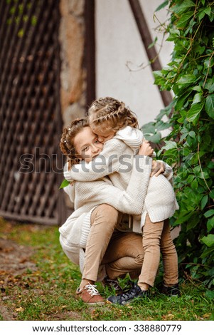 Two charming sisters in the same curly beige knitted sweater embracing and kissing on the background of a wooden house and green grapes in autumn