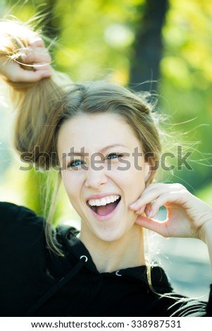 Portrait closeup of laughing pretty blond girl with long hair and big joyful smile taking hair in hands posing outside on blurred streetscape background, vertical picture