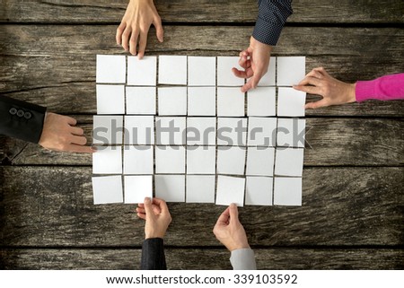 Top view of six hands, male and female, assembling a collage of blank white cards over textured wooden rustic boards.
