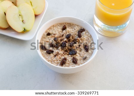 A simple bowl of raisin and cinnamon oatmeal, a glass of orange juice, and a sliced apple on a white tile background. This delicious scene was lit by the natural, early morning light through a window.