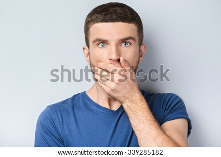 Portrait of handsome caucasian man standing on grey background. Young man covering his mouth with hand and looking at camera