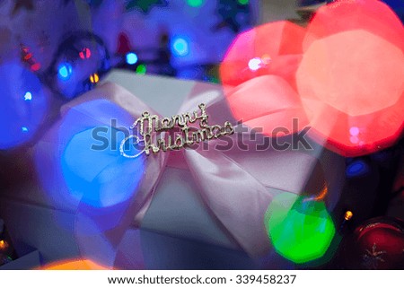 Image gift that says "Merry Christmas" in the rays blurred festive lights. The picture is a Christmas present on background of colored lights garlands.