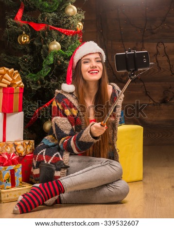 Beautiful girl sitting on the floor near New Year tree and making selfies. Happy lady in Santa hat and stripped socks smiling for the camera.