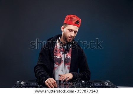 Closeup portrait of confident DJ with stylish red cap and headphones on neck mixing music on mixer looking up while standing isolated on dark background