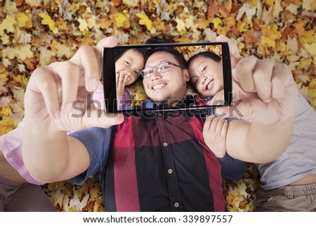 Portrait of young man and his children taking selfie photo with smartphone while lying on the autumn leaves