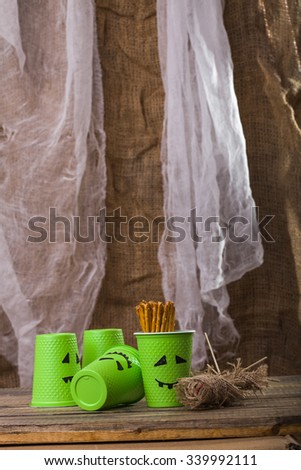 Photo still life green disposable cups with Halloween ghost scary faces smiles one filled with straws and rag voodoo doll on wooden table over blurred rustic background, vertical picture 