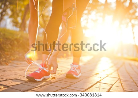 Young beautiful girl athlete with armband and earphones listening to music and tying laces during training with sunset and sunbeam on background. Copy space. Bright and warm photo