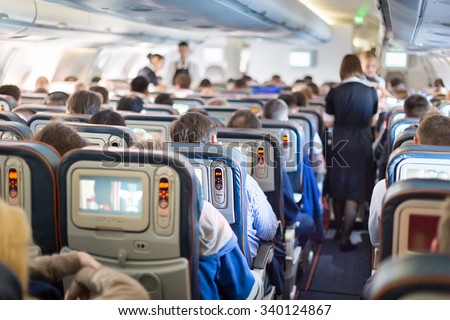 Interior of large passengers airplane with people on seats and stewardess in uniform walking the aisle.  Royalty-Free Stock Photo #340124867