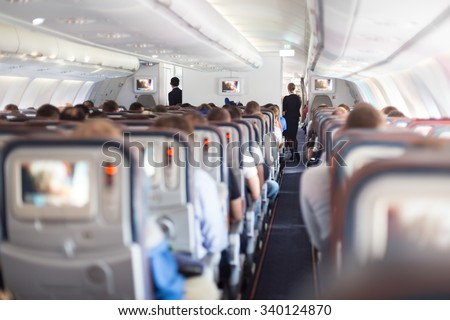 Interior of large passengers airplane with people on seats and stewardess in uniform walking the aisle.  Royalty-Free Stock Photo #340124870